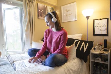 Student on a bed with headphones looking at a laptop