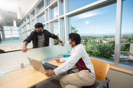 Two students in library wearing masks and talking with laptop between them