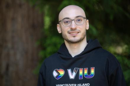 VIU grad and current employee Rayan Zeineddine wearing a VIU hoodie and glasses standing next to a tree outside and smiling at the camera